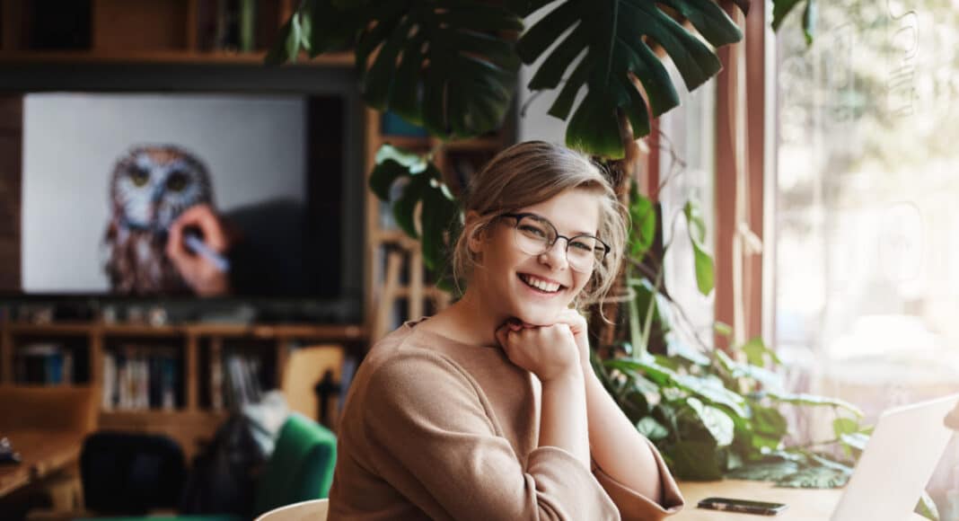 Girl waited for friend in cafe, smiling and saying hi, being glad to finally meet. Charming and carefree european female with fair hair in glasses, sitting near window and laptop, leaning on hands.